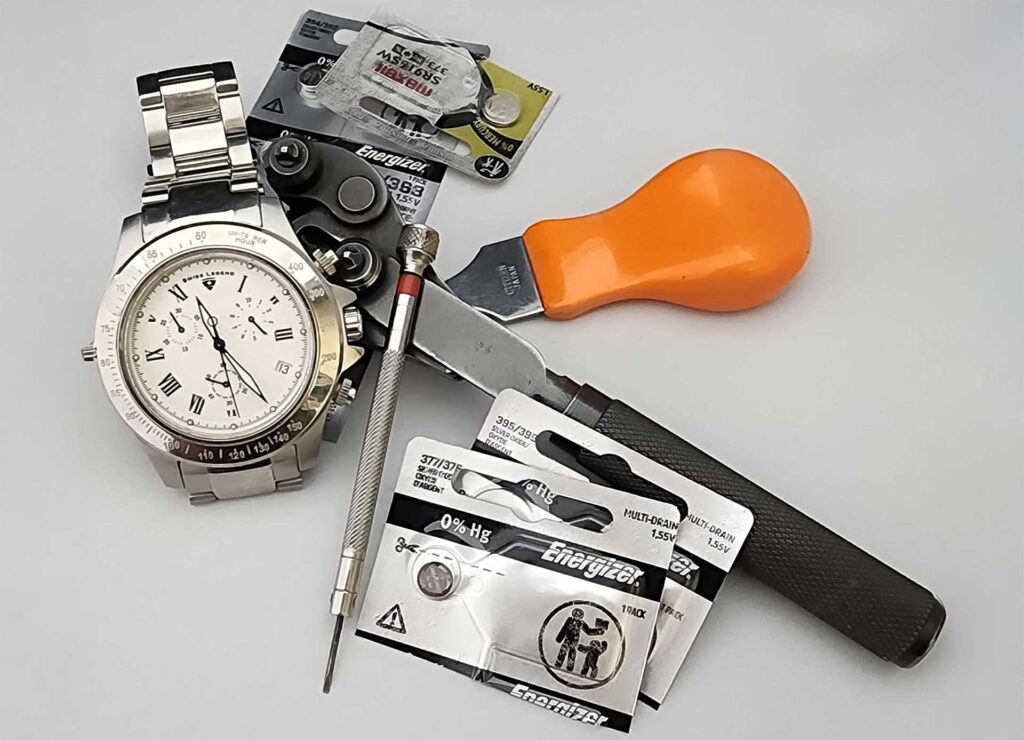 Watch repair tools and batteries arranged around a silver wristwatch on a white background.
